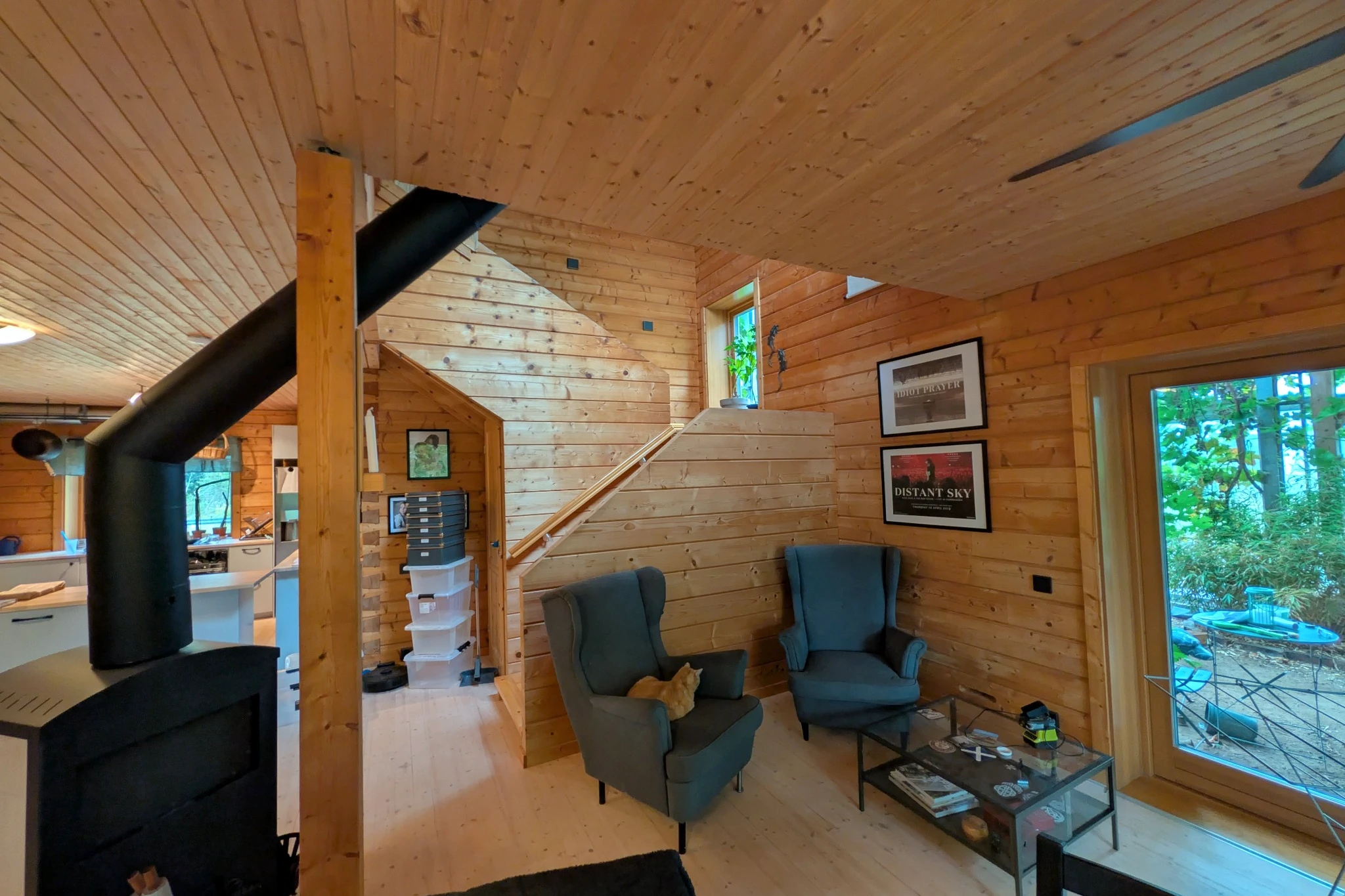 Timber-lined living room with a wood-burning stove, two armchairs and a glass door opening towards the greenhouse.